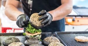 At a creative food station for a corporate event, a person assembles a burger with black sesame seed buns, expertly placing fresh arugula on top of the sizzling patty at the grill station.