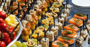 A variety of colorful appetizers on skewers are elegantly displayed on a table, perfect for a corporate event, featuring salmon, shrimp, and fruit. Plates are stacked in the background, offering creative food station ideas that add flair to any professional gathering.