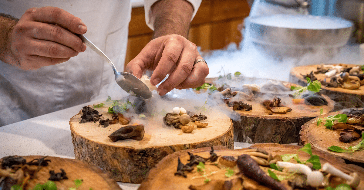 In the heart of Salt Lake City, a chef crafts gourmet mushroom dishes, artistically plating them on wooden boards amid the misty allure of liquid nitrogen vapor. Perfect for any upscale catering service seeking a touch of culinary theater.