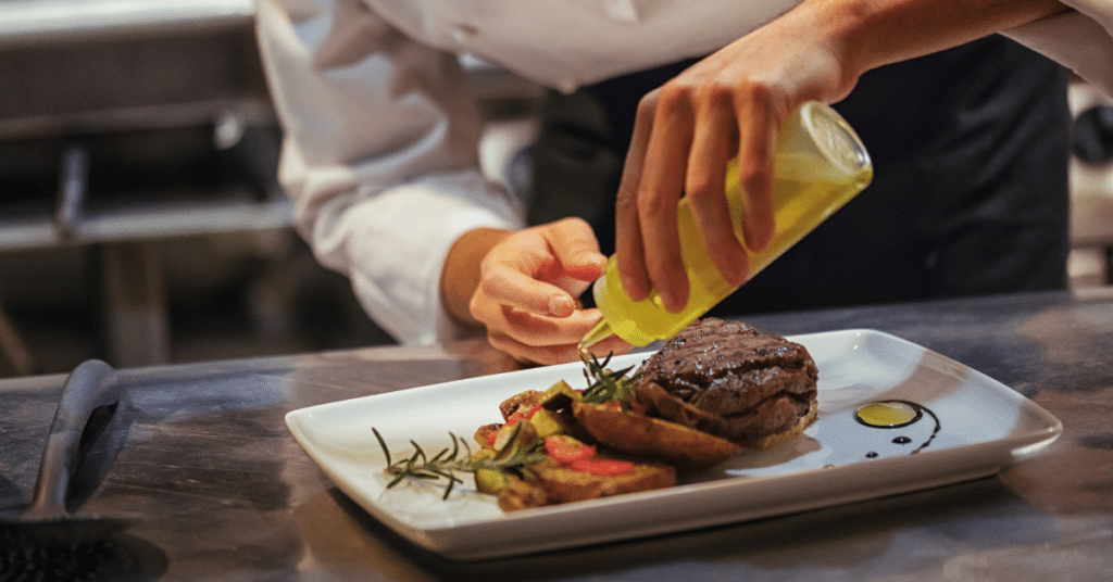 In a Salt Lake City kitchen, the chef expertly plates a dish featuring steak, vegetables, and sauce. With precision, he uses a squeeze bottle of yellow dressing on the white rectangular plate, embodying the artistry of catering service.