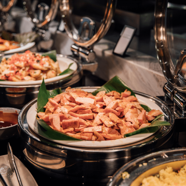 A serving dish lined with banana leaves holds sliced pieces of luncheon meat at a lunch catering event in Orem, Utah, with other food trays in the background.