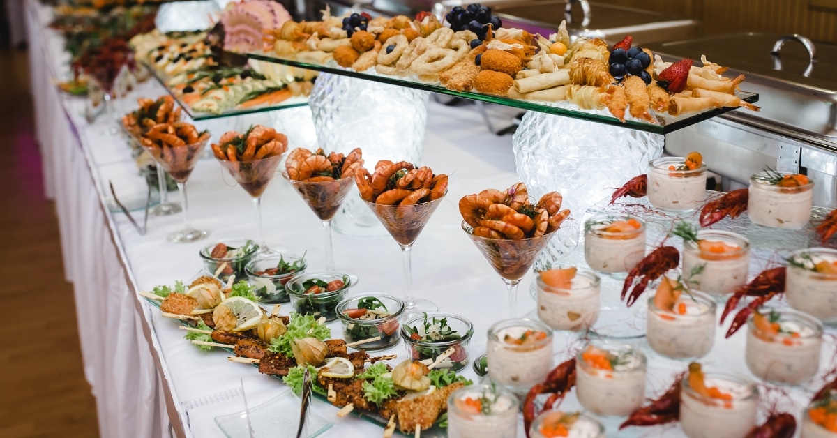 A buffet table showcasing catering delights with assorted appetizers, like shrimp cocktails in glasses and meat skewers, pastries, and seafood in small jars. Perfect for seasonal wedding menus in Utah, it's elegantly decorated with grapes on a pristine white tablecloth.
