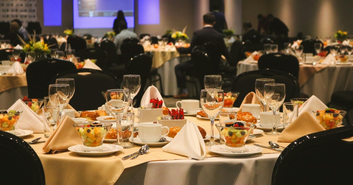 Round banquet tables set with fruit bowls, pastries, and glassware in a dimly lit event hall, with people seated in the background and a projection screen—perfect for holiday catering events in Orem Utah.